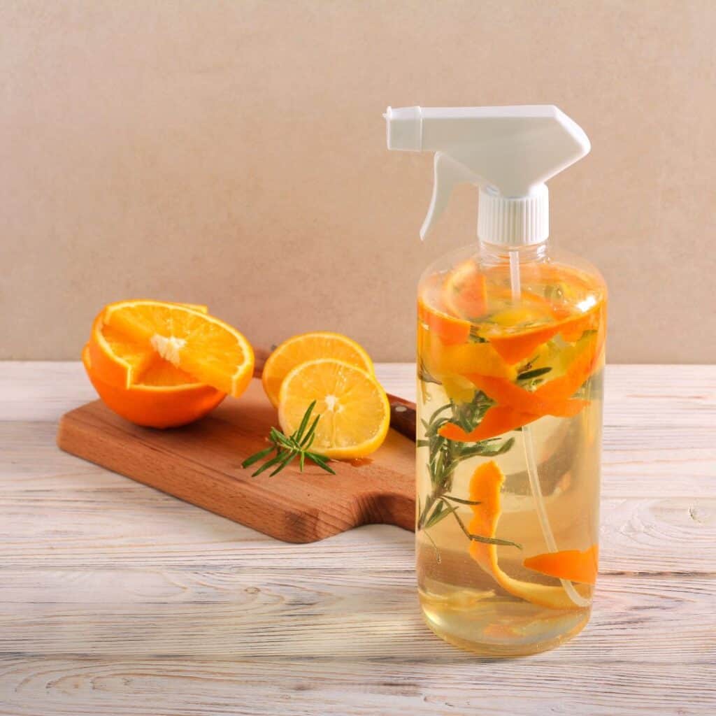 clear spray bottle with white spray top filled with liquid and oranges and sliced oranges on cutting board behind the spray bottle