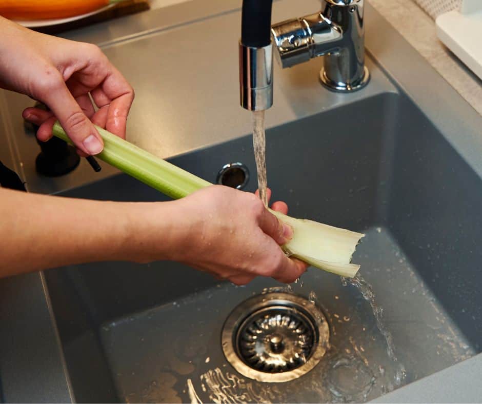 one stalk of green celery being held by woman's hands under running water from facet for how to clean celery post